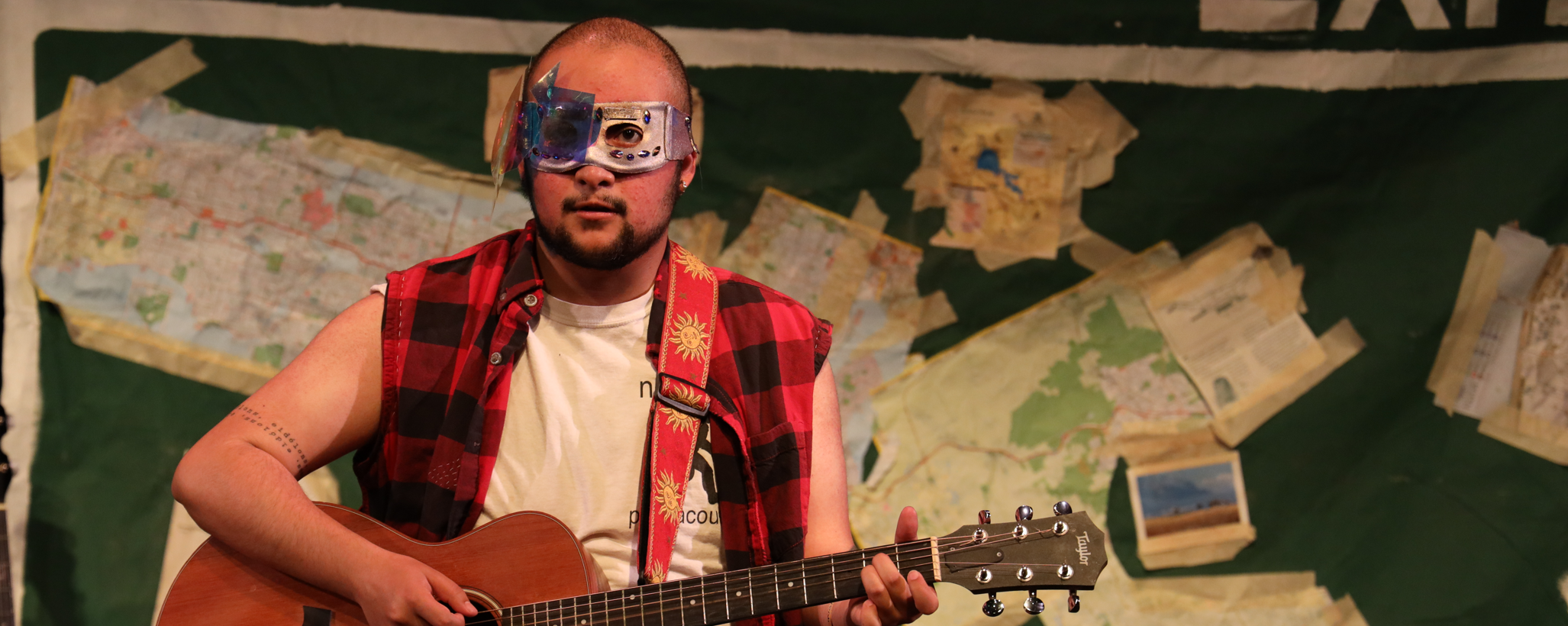 Walden, a brown man in a half-mask, plays his guitar while looking directly ahead.